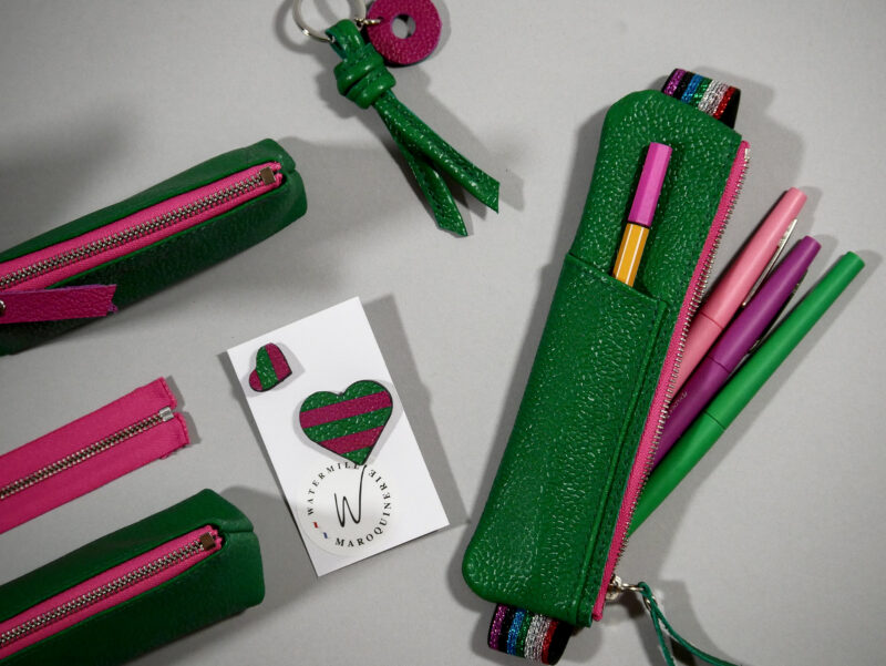 trousse élastique en cuir grainé vert émeraude, zip contrasté fushia et avec compartiment avant pour accès rapide , élastique arc en ciel, présenté avec accessoires coordonnés: pin's, porte clefs, trousse berlingot