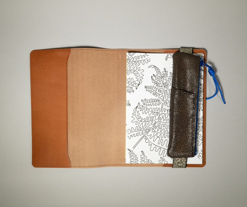 Carnet cuir format A5 — couverture de carnet et protège agenda en cuir lisse camel, présenté ouvert avec un carnet noir et blanc glissé dans l'un des deux emplacements intérieurs, maintenu fermé par la Trousse élastique en croûte de cuir noir pailleté.