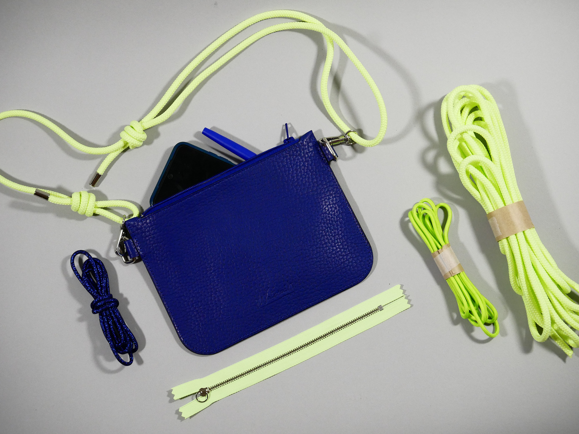 la Nomade: pochette en cuir grainé bleu roi avec sangle en paracord jaune fluo ajustable grâce à des noeuds présentée avec des Paracord et un zip fluo