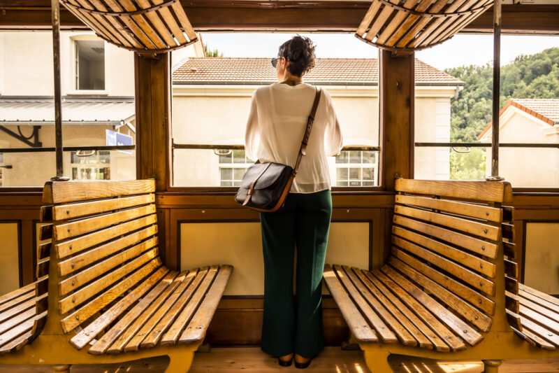 Besace en cuir noir fermée par rabat et pontet en cuir contrasté camel, porté par mannequin dans train vintage wagon bois