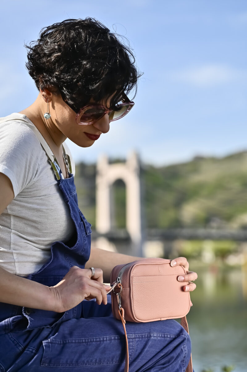 sac camera case en cuir grainé rose poudré, de forme rectangulaire, avec poche avant plaquée porté prêt du corps comme une banane grâce à sa sangle en corde, mannequin femme , photo en extérieur sur fond de port de plaisance