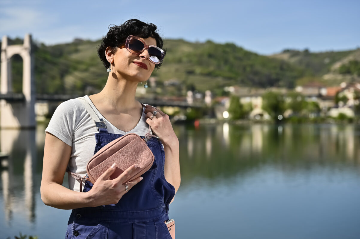 sac camera case en cuir grainé rose poudré, de forme rectangulaire, avec poche avant plaquée porté prêt du corps comme une banane grâce à sa sangle en corde, mannequin femme , photo en extérieur sur fond de port de plaisance