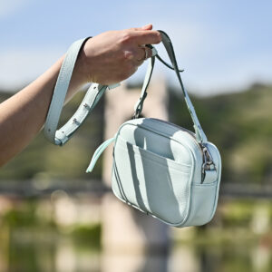 Sac à main camera case, en cuir de couleur cyan, avec bandoulière et poche avant porté à la main sur fond de port de plaisance