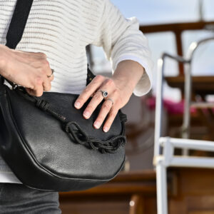Sac demi-lune en cuir noir avec sangles en corde et cuir, porté à l'épaule par le mannequin, devant un bateau