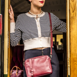 Sac Reporter en cuir grainé bordeaux, bandoulière réglable laiton vieilli, porté en bandoulière par une femme sur le quai d'un train vintage.