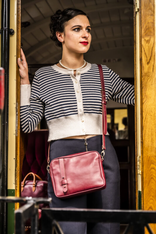 Sac Reporter en cuir grainé bordeaux, bandoulière réglable laiton vieilli, porté en bandoulière par une femme sur le quai d'un train vintage.
