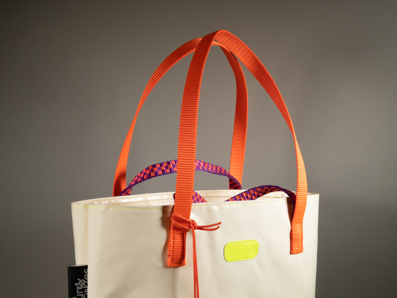 Détails des sangles : Tote bag en bâche blanche upcyclée, double anse — sangle plate orange fluo et corde paracord violet et orange tressé — logo Watermill jaune fluo, avec porte-clés cloche en bâche transparente attaché par une cordelette paracord orange. Étiquette Pures Folies visible sur le côté.
