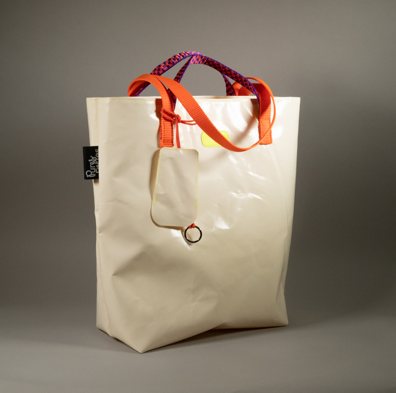 Tote bag en bâche blanche upcyclée, double anse — sangle plate orange fluo et corde paracord violet et orange tressé — logo Watermill jaune fluo, avec porte-clés cloche en bâche transparente attaché par une cordelette paracord orange. Étiquette Pures Folies visible sur le côté.