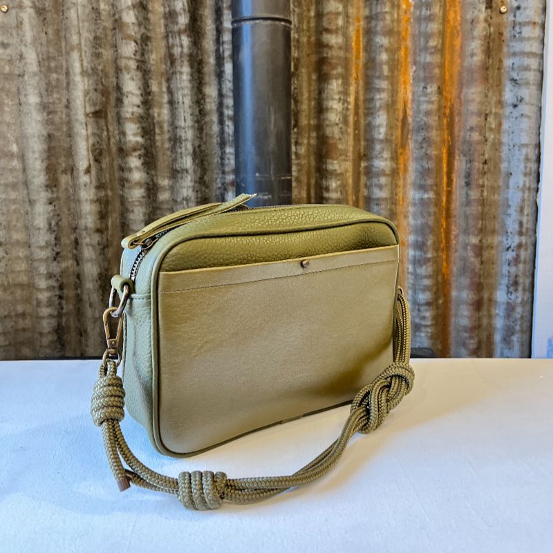Sac Camera Case M Watermill bicolore cuir grainé kaki et poche avant plaquée surpiquée vert mordoré, bouclerie et zip métal laiton vieilli, présenté avec sa sangle paracord coordonnée nouée façon macramé, présenté sur fond de métal brut.