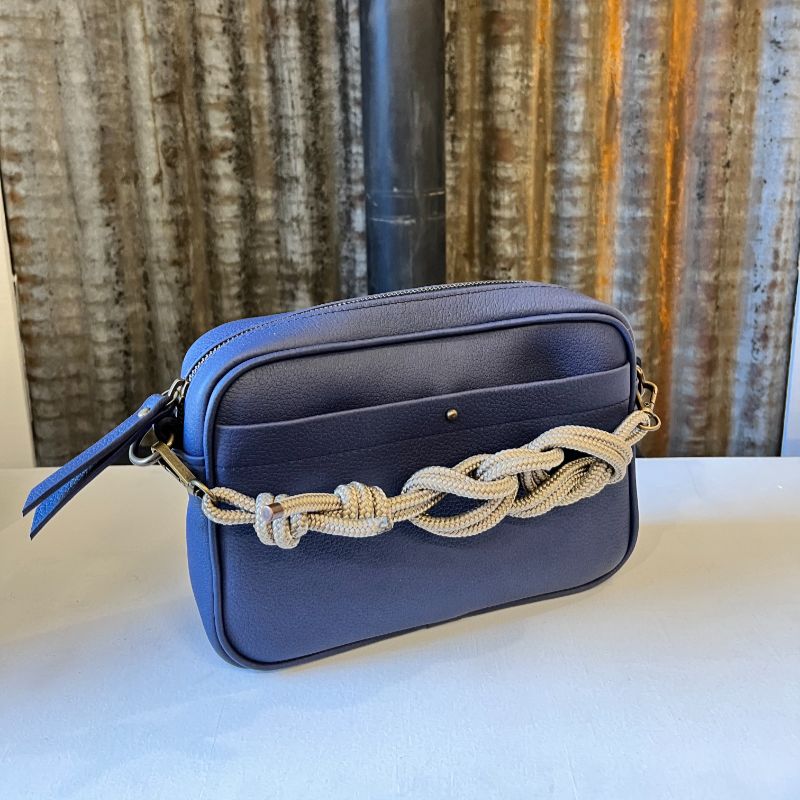Sac Camera Case M Watermill bicolore cuir grainé bleu marine et poche avant plaquée surpiquée bouclerie et zip métal laiton vieilli, présenté avec sa sangle paracord coordonnée nouée façon macramé, présenté sur fond de métal brut.