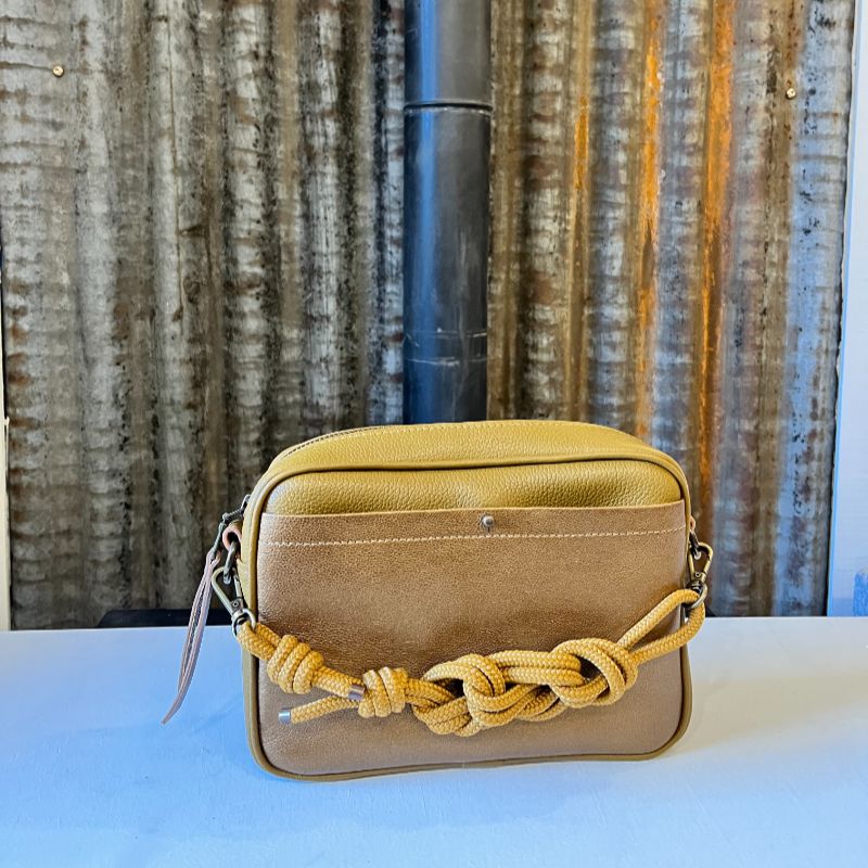 Sac Camera Case M Watermill bicolore cuir grainé comice et poche avant plaquée surpiquée doréé, bouclerie et zip métal laiton vieilli, présenté avec sa sangle paracord coordonnée nouée façon macramé, présenté sur fond de métal brut.