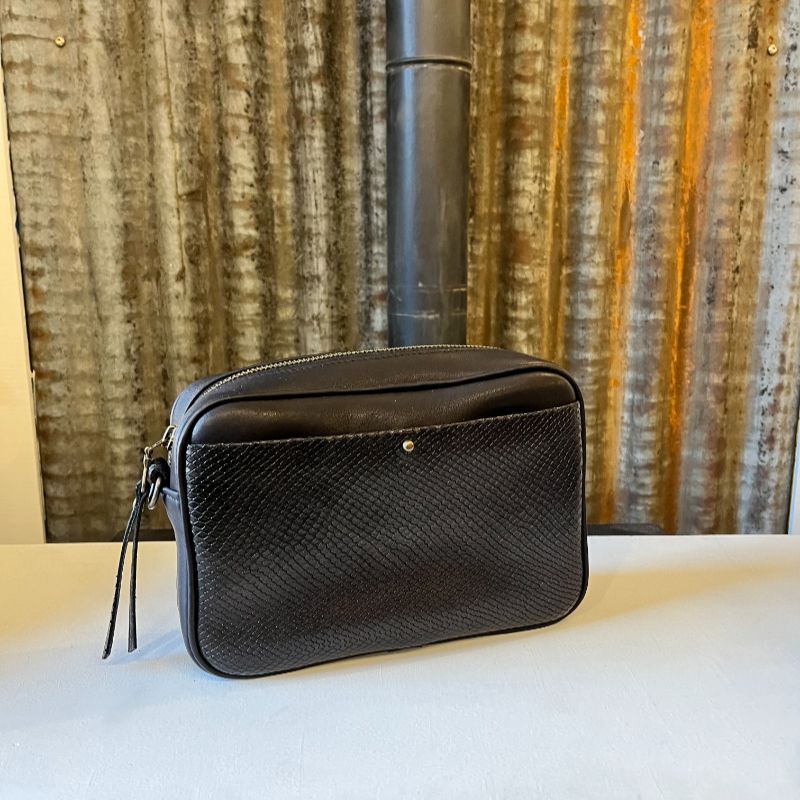 Sac Camera Case M Watermill bicolore cuir lisse noir et poche avant plaquée surpiquée noire façon écailles, bouclerie et zip métal laiton vieilli, présenté avec sa sangle paracord coordonnée nouée façon macramé, présenté sur fond de métal brut.