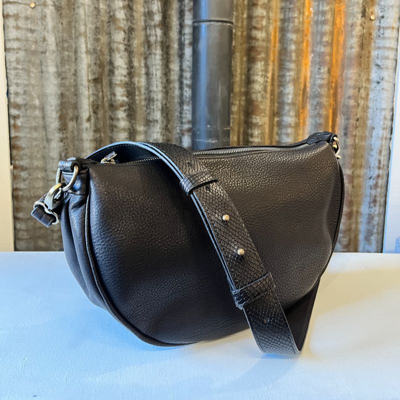 Sac demi-lune en cuir noir lisse et avec sangles en cuir large en cuir contrasté noir façon écailles, présenté devant une tôle d'atelier